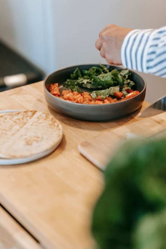 Crop unrecognizable person adding spices into plate with delicious food on table of modern kitchen